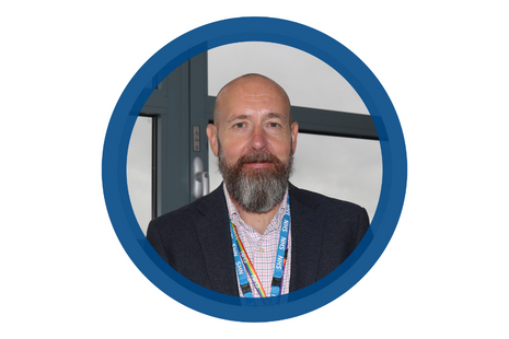 Pictured Lee Neary - bald head with dark brown and grey pointed beard wearing a navy suit jacket and white with pink and dark blue check shirt (relaxed open fit) with NHS lanyard.